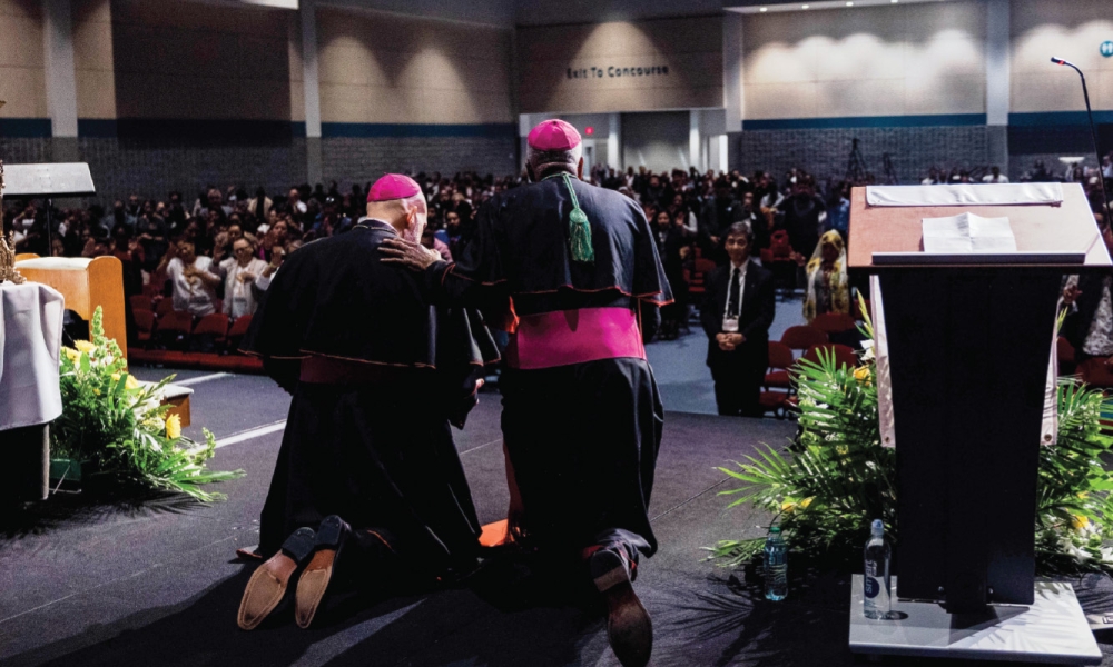 Bishop Jacque Fabre-Jeune kneeling on a stage before an audience.